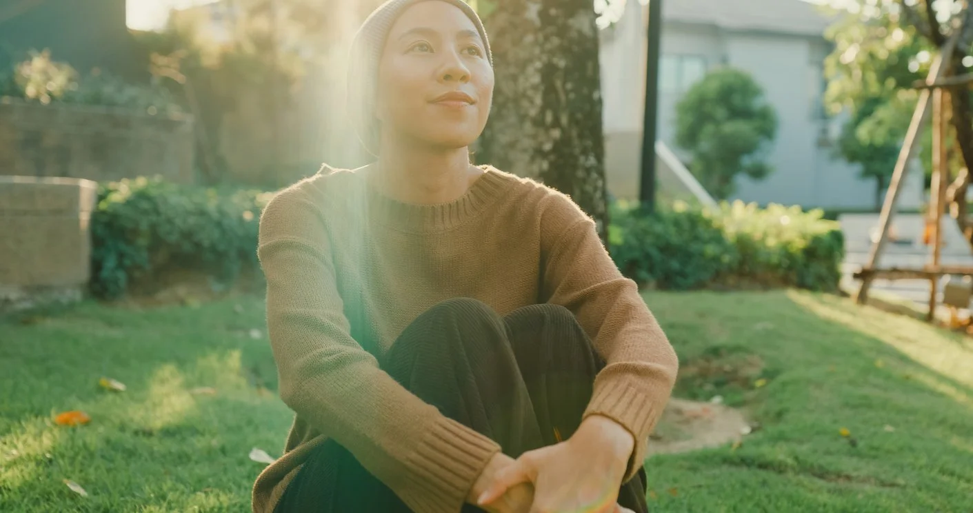 A woman sitting on the grass in a park or backyard during the daytime, with sunlight shining through trees in the background, wearing a beige sweater and a beanie.