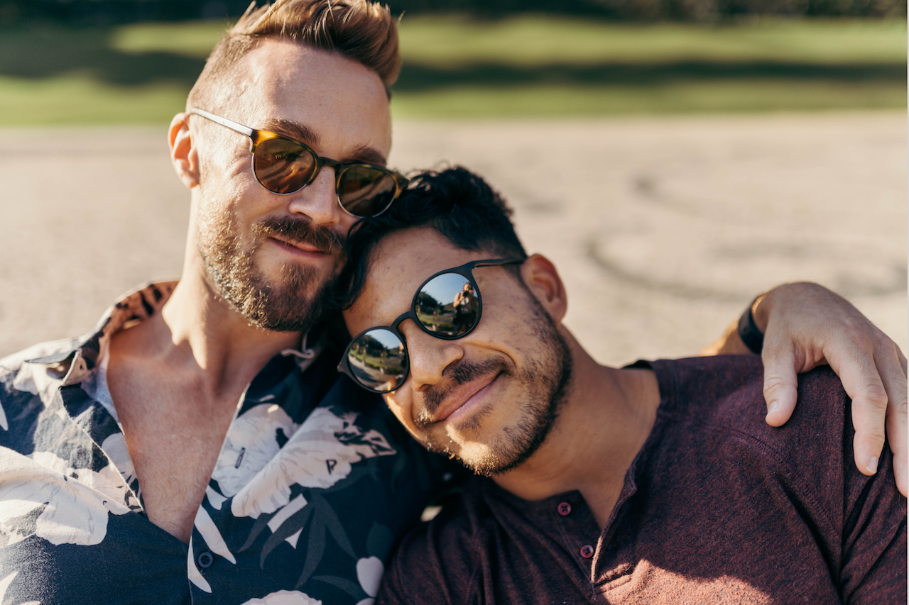 Two men with sunglasses smiling and hugging at the beach on a sunny day.