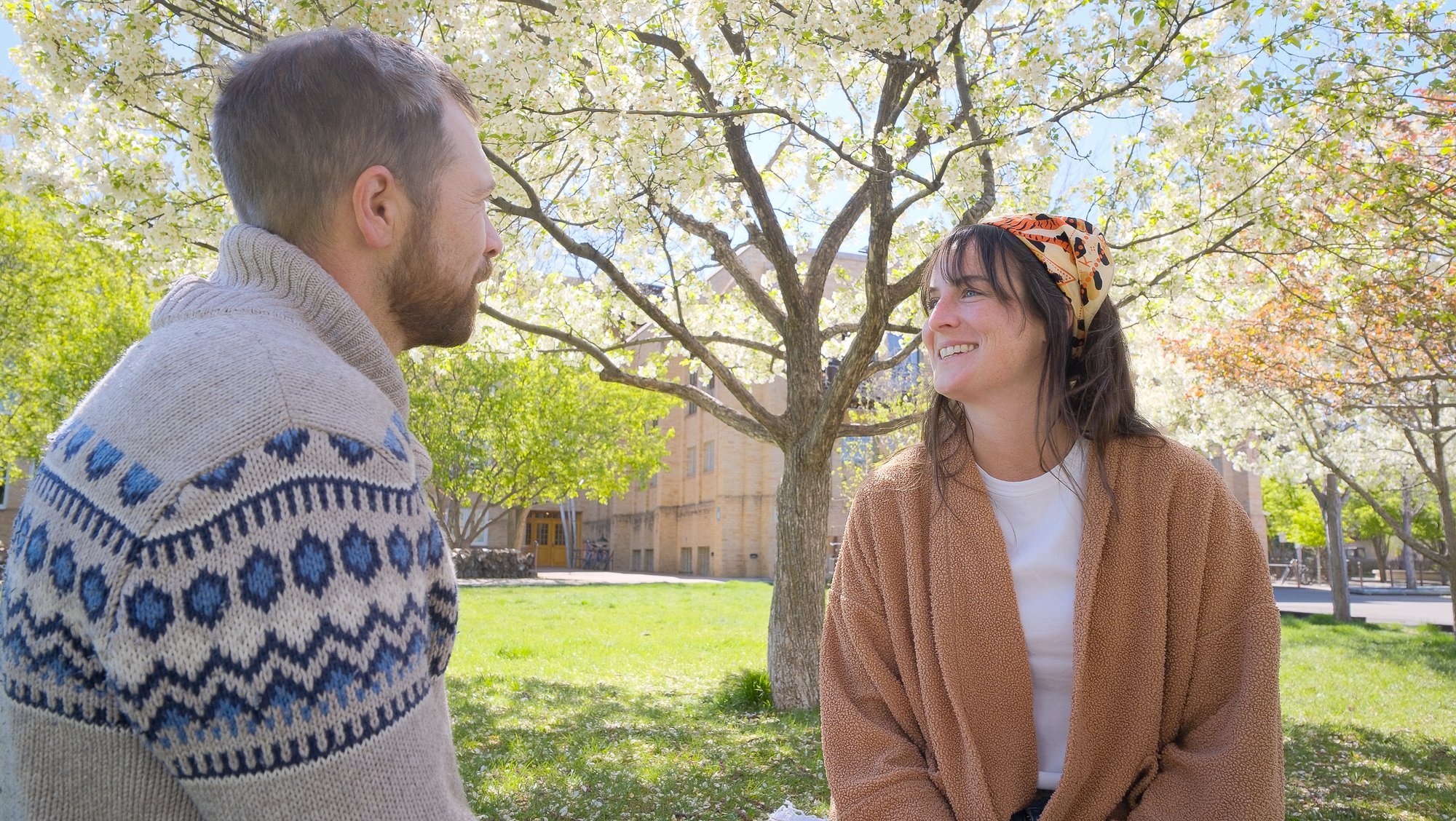 A man and woman sitting outdoors under blossoming trees, smiling and engaging in conversation.
