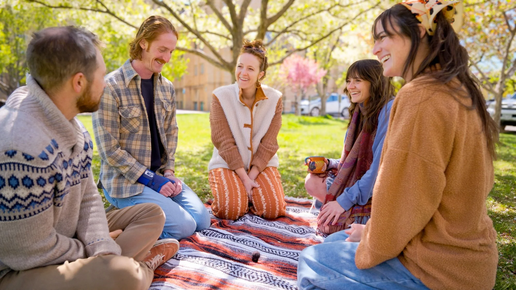 Group of six young adults sitting on a blanket outdoors, laughing and talking on a sunny spring day with trees and buildings in the background.