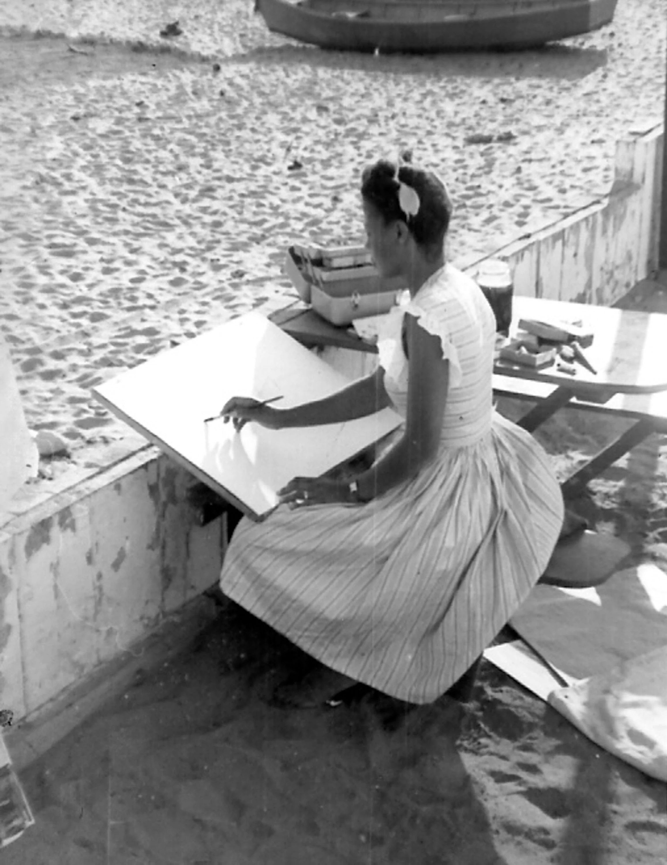 A woman with her hair tied back, wearing a dress with ruffled sleeves, is seated at a wooden table on a sandy beach, writing or drawing on a paper pad.