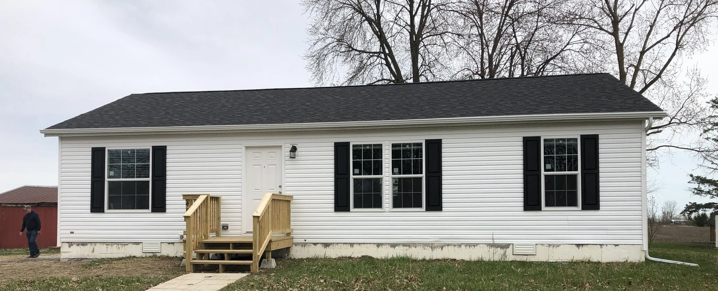 Newly constructed white house with black shutters and a small wooden front porch, situated on a grassy lot with mature trees in the background.