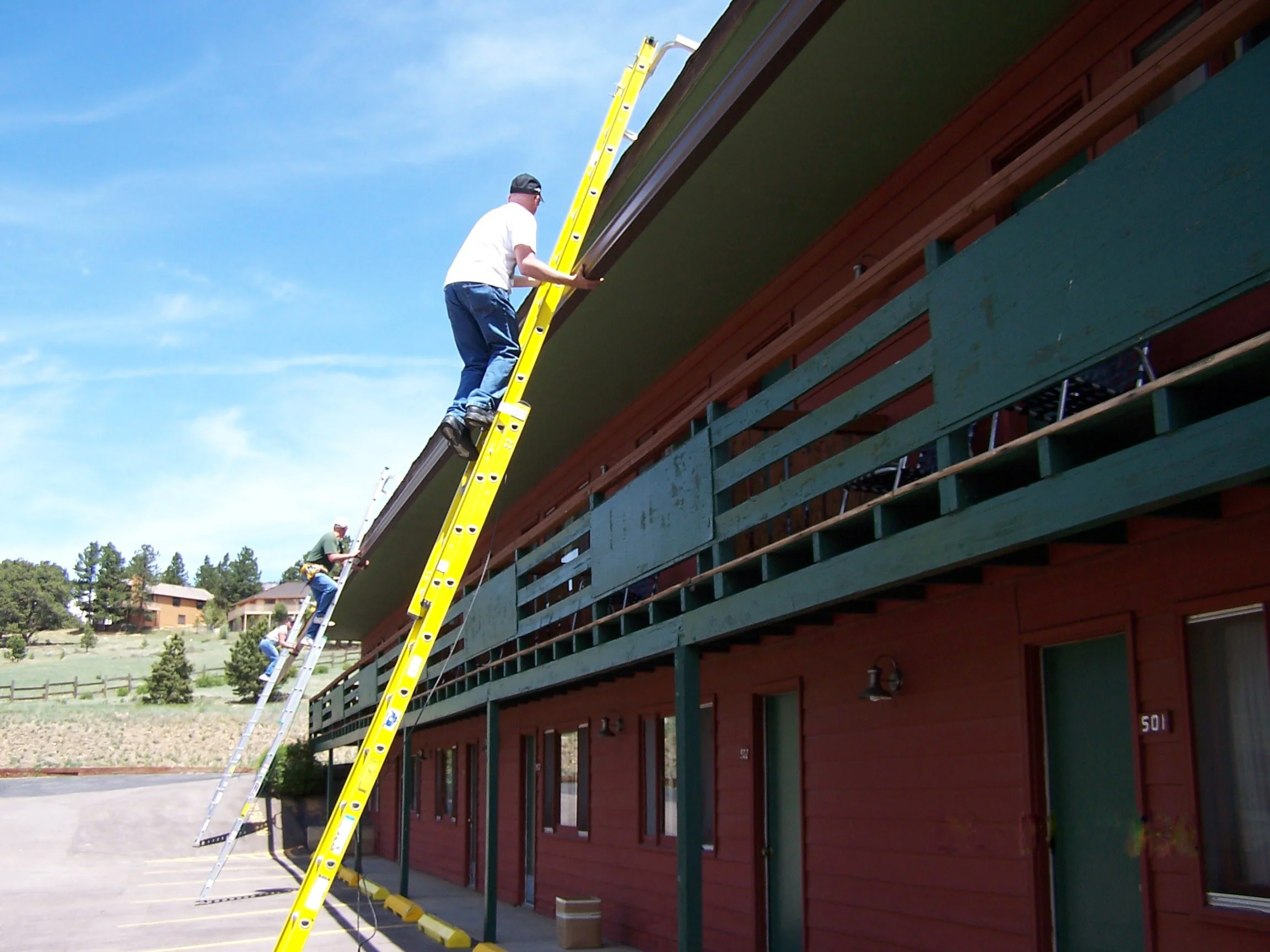 Two men are working on the roof of a red two-story building. They are positioned on ladders, one on a yellow ladder and the other on a silver ladder, installing or repairing something along the edge of the roof.