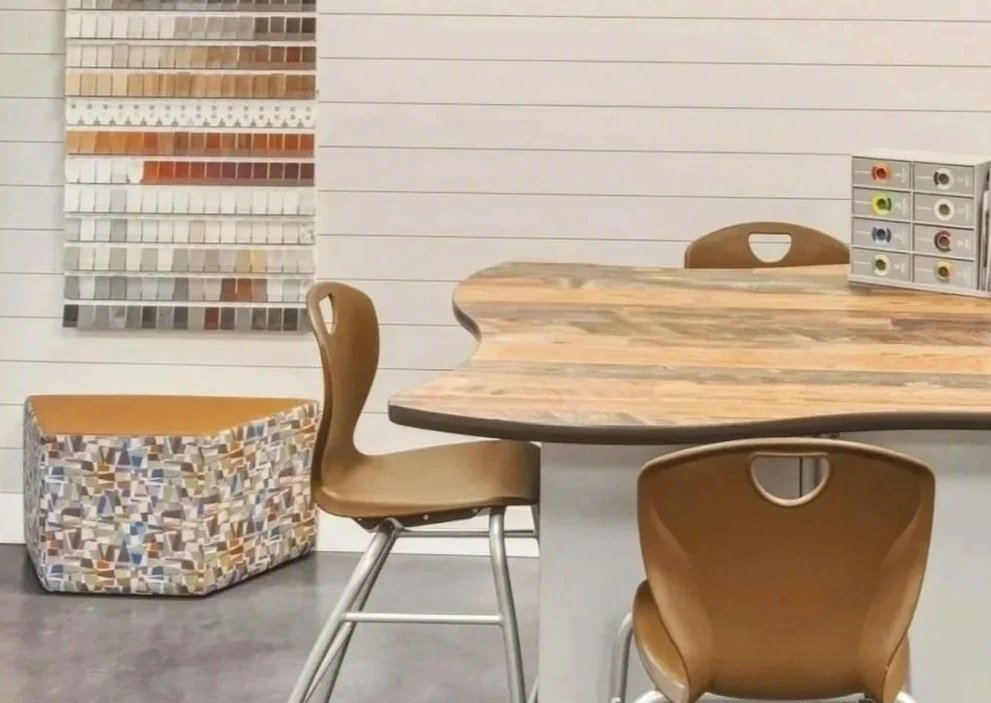 A wooden table with three brown chairs in a room with a white wall. On the wall, there is a display of various tile samples. To the left, a fabric-covered storage box with a geometric pattern sits on the floor. On the right, a small drawer unit holds colored ink or paint bottles.