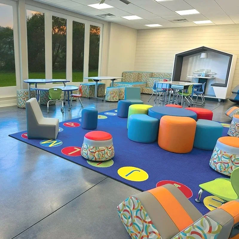 Colorful children's playroom with soft seating, small tables and chairs, a game rug with numbers and letters, and large windows showing an outdoor view.