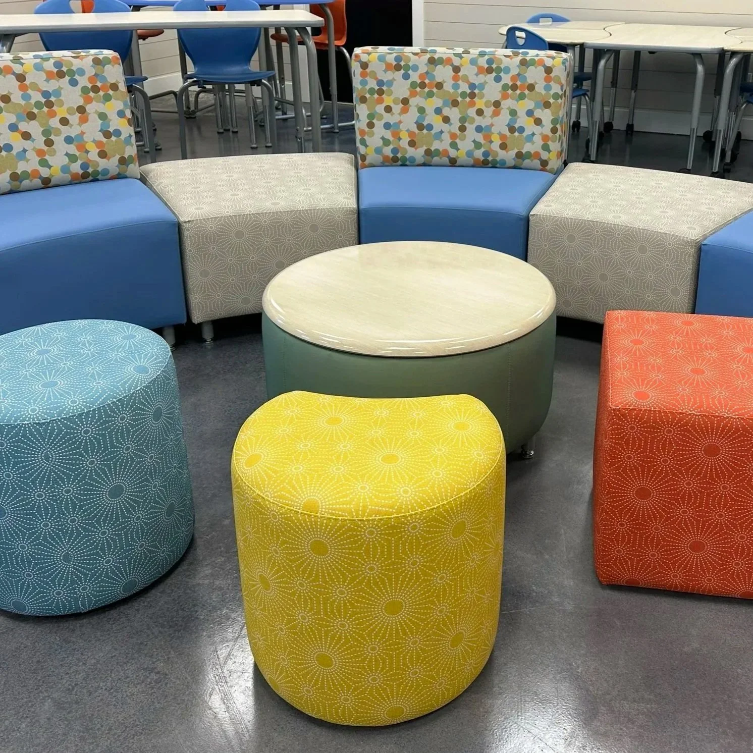 Colorful seating furniture including a round beige table, yellow, orange, and blue ottomans with geometric patterns, and a modular sofa with patterned and plain cushions in a modern indoor space.