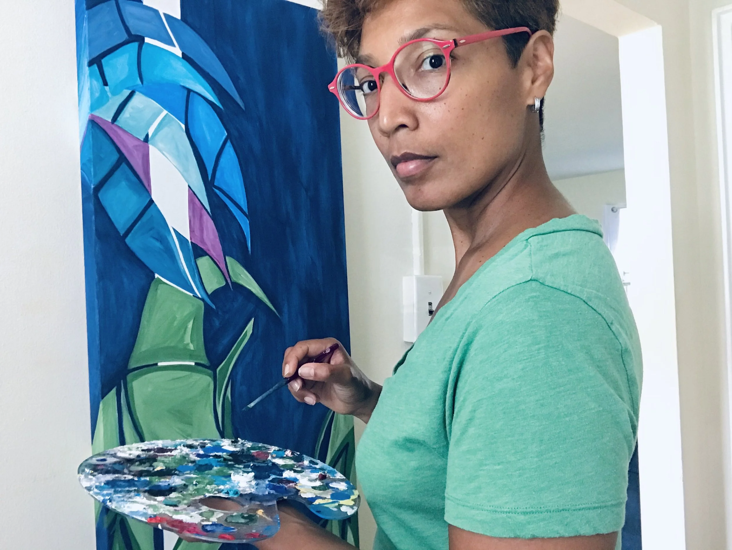 An artist with short hair, pink glasses, and earrings holding a paint palette and brush, working on a colorful abstract painting of leaves on a blue background.