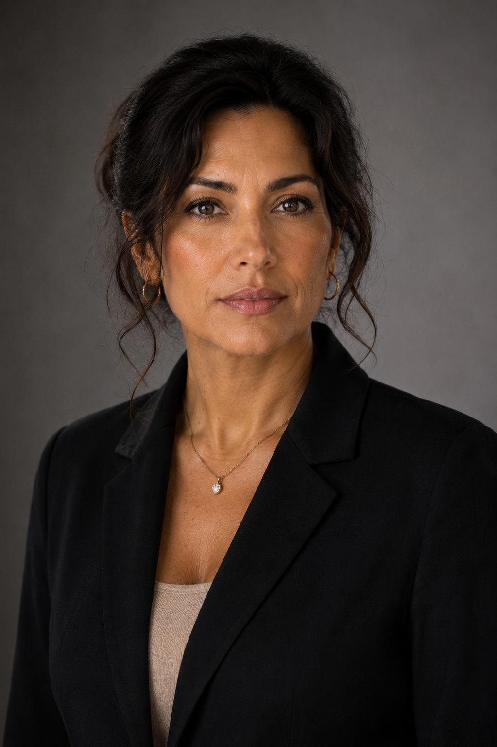 Headshot of a middle-aged woman with dark, wavy hair, wearing a black blazer, beige top, small hoop earrings, and a delicate necklace with a round pendant, against a gray background.