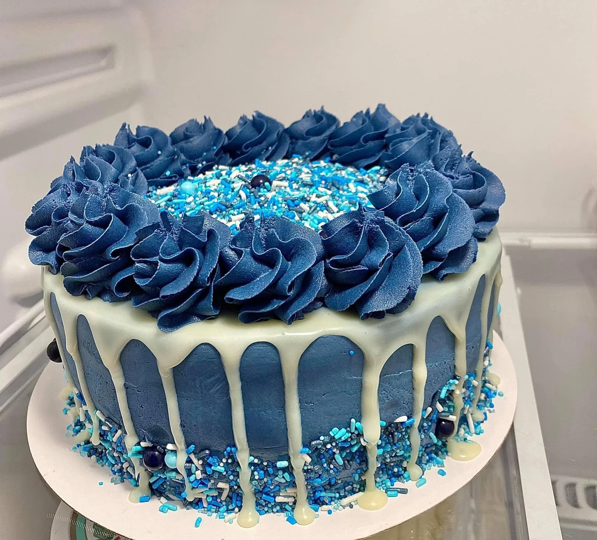 A round cake decorated with blue and white icing, white chocolate drip, blue and white sprinkles, and blue frosting rosettes on top.