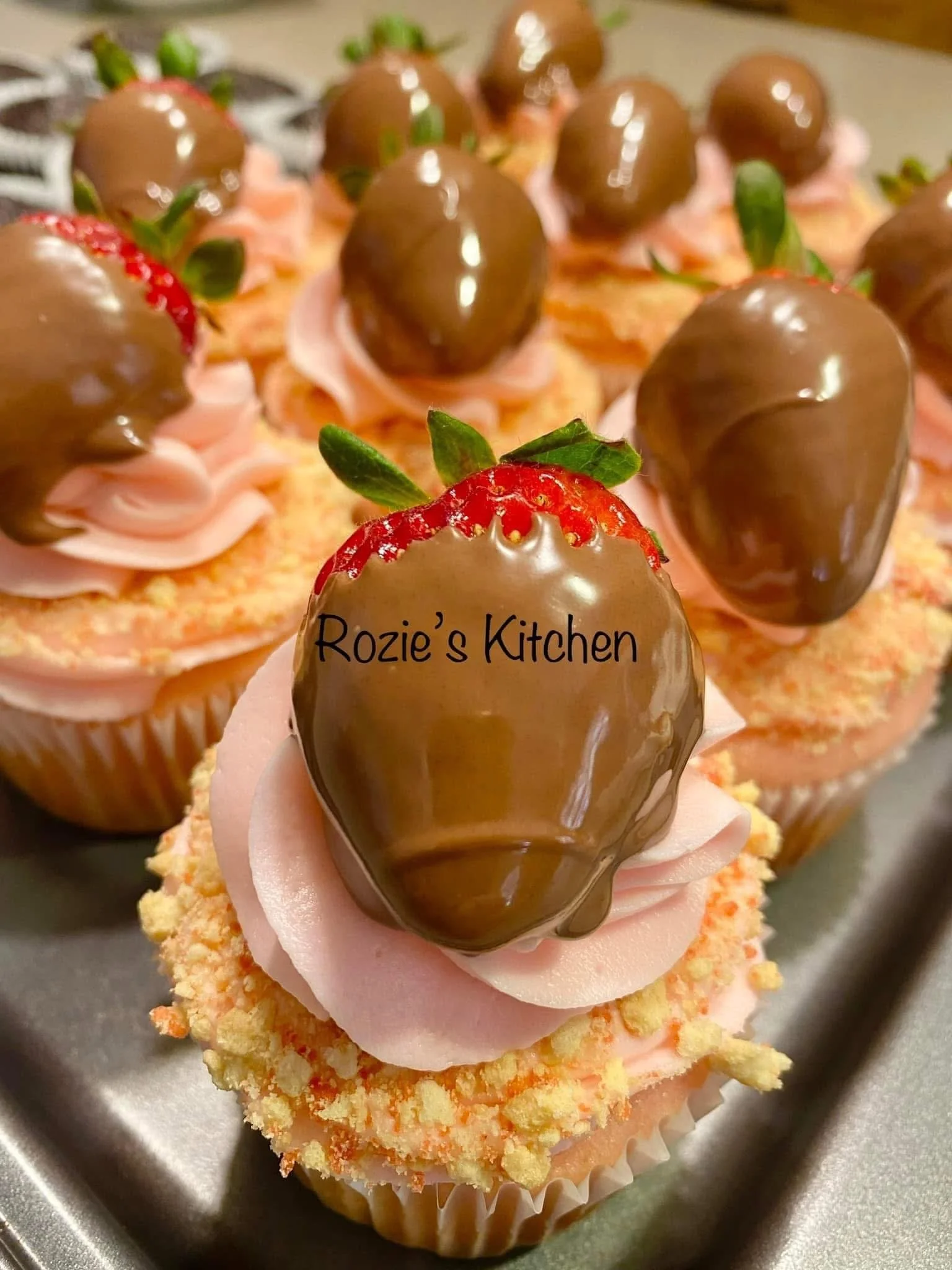 Close-up of decorated strawberry cupcakes with chocolate-dipped strawberries on top. One cupcake has the text 'Rozie's Kitchen' written on the chocolate-covered strawberry.