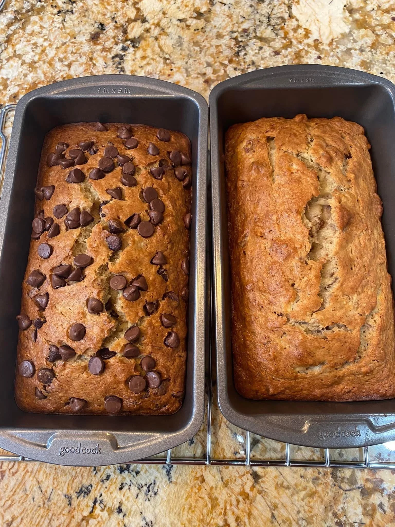 Two loaves of baked bread in grey baking pans on a wire rack, placed on a speckled beige and brown countertop. The loaf on the left is topped with chocolate chips; the loaf on the right is plain.