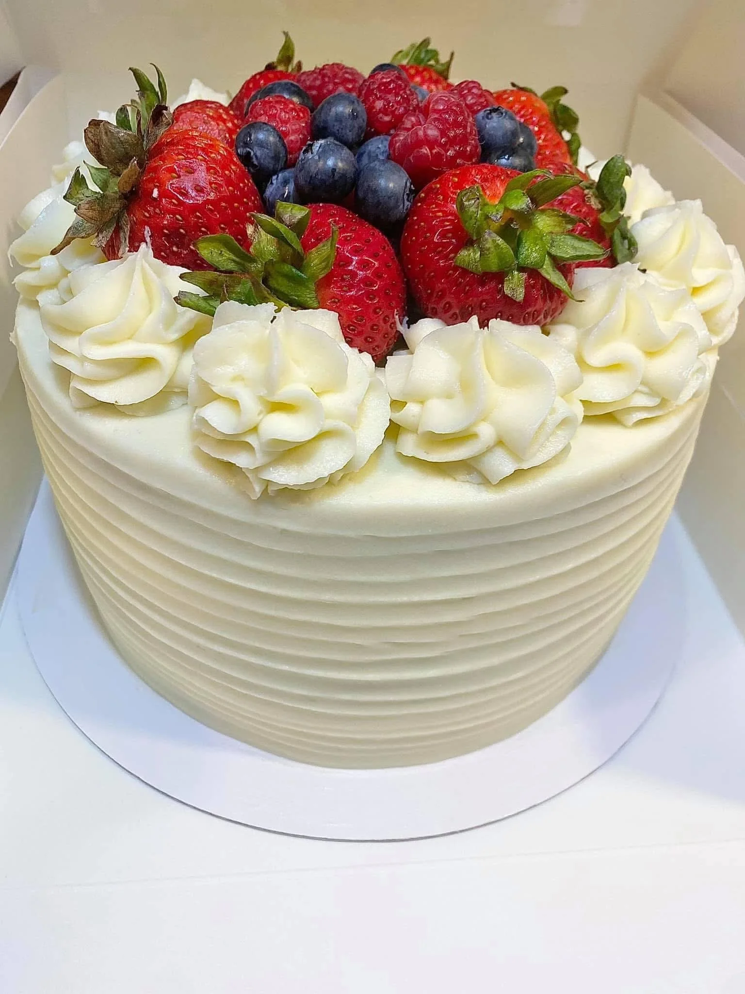 A heart-shaped cake decorated with white frosting, topped with strawberries, blueberries, and raspberries, with piped whipped cream around the top edge.