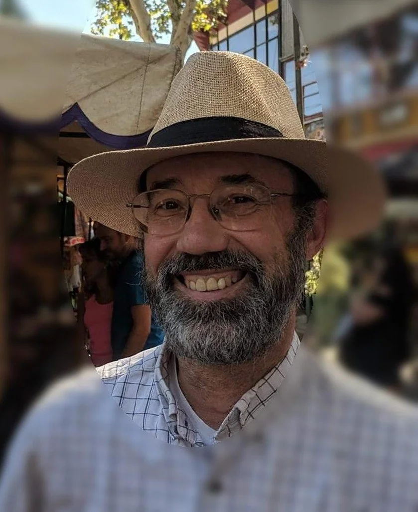 Man with glasses, gray beard, and mustache smiling, wearing a straw hat with a black band, white checkered shirt, outdoors with blurred people and buildings in background.