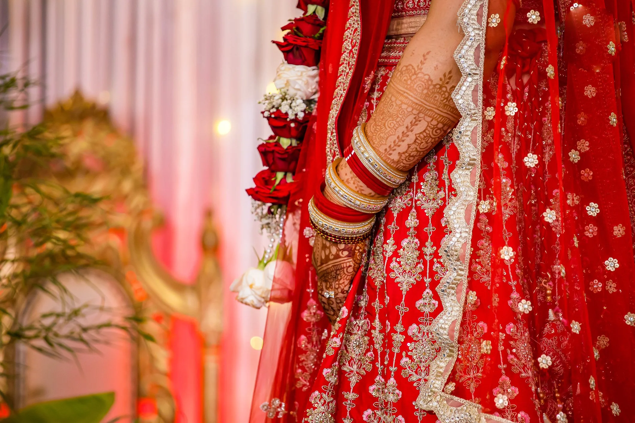 Close-up of a woman in traditional red bridal attire with intricate embroidery, henna designs on her arm, and multiple gold and red bangles at a wedding.