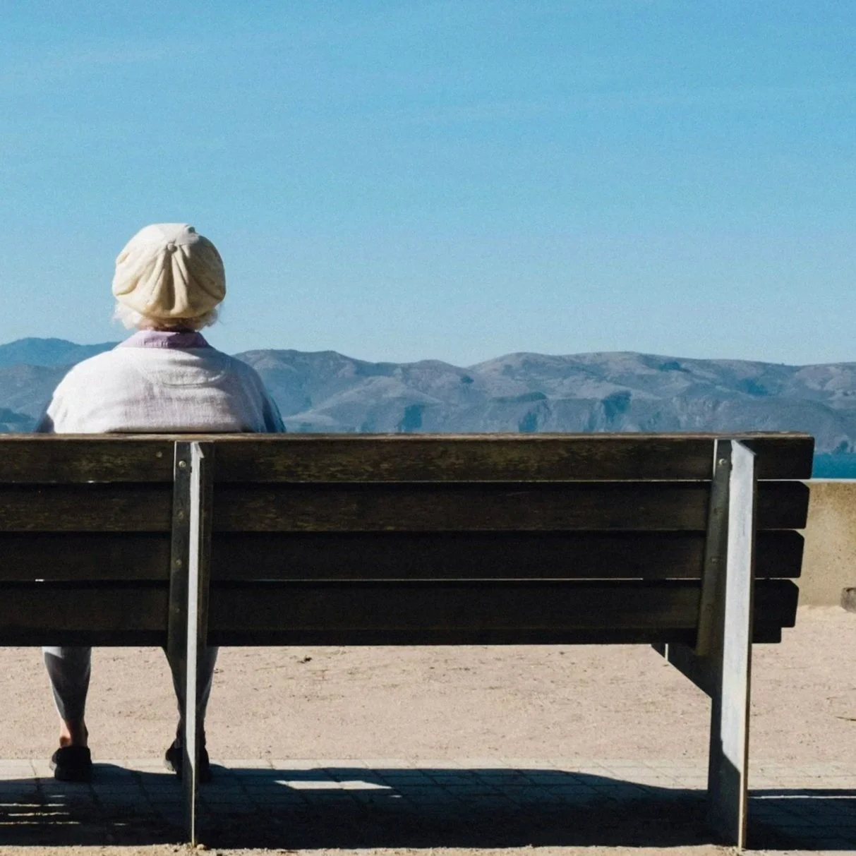 Person sitting on a wooden bench overlooking mountains under a clear blue sky.