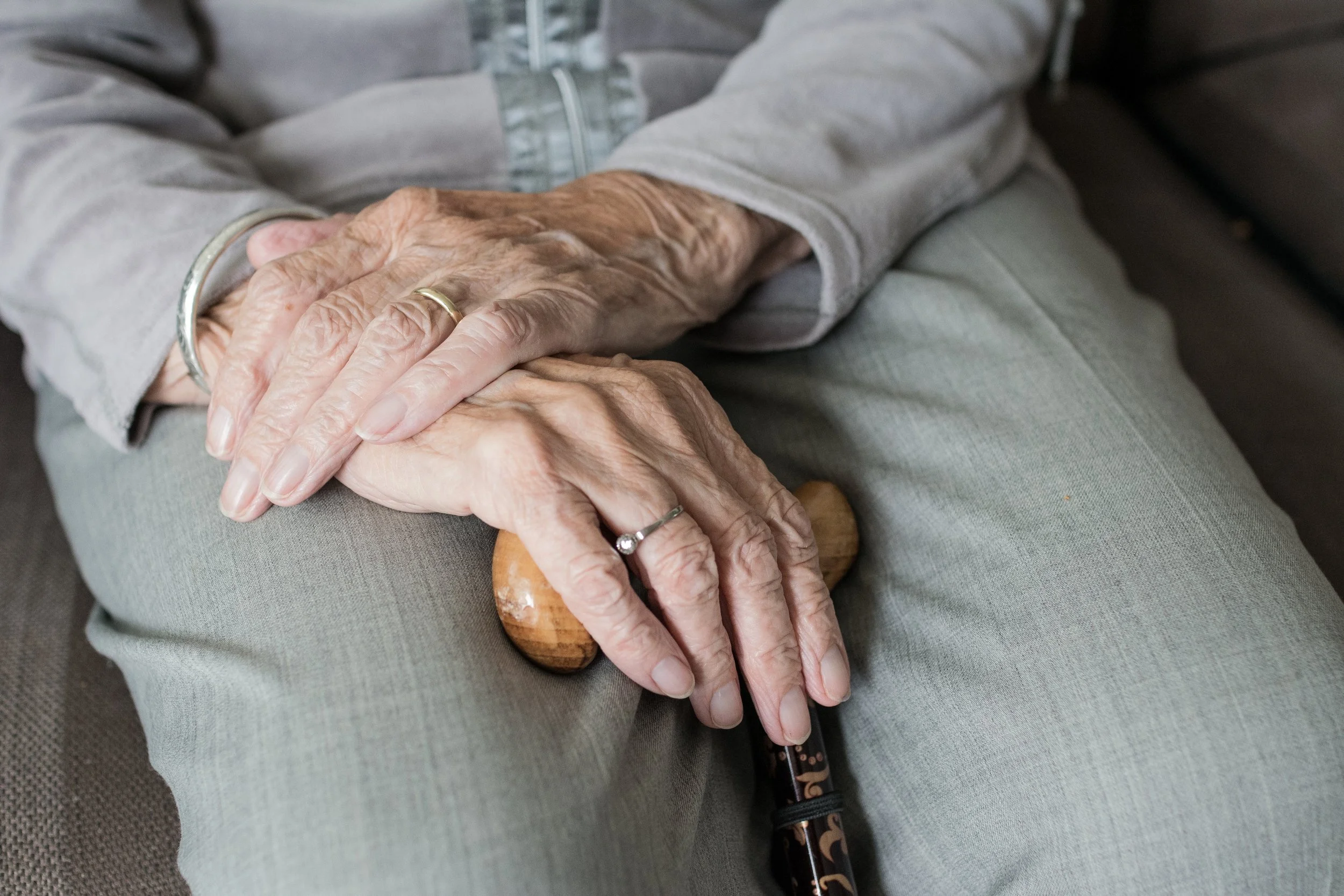 Close-up of elderly hands, one with a gold ring, clasping a cane with a carved wooden handle, resting on lap.