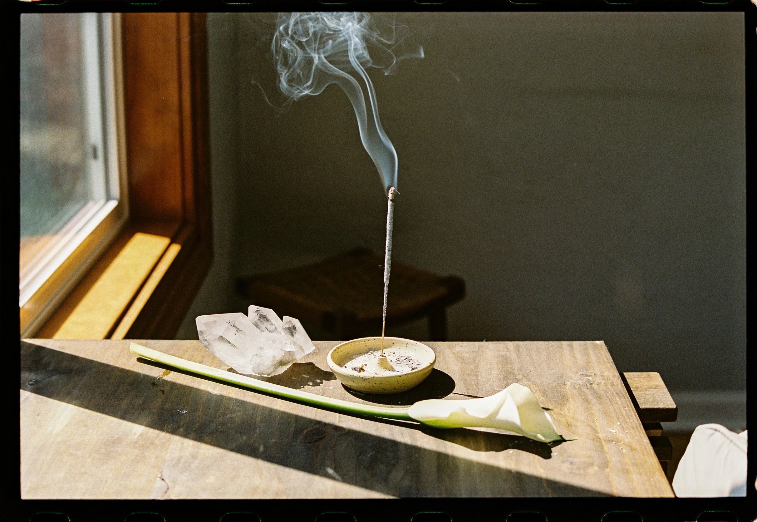 A lit incense stick producing smoke in a bowl on a table, with a leaf and ice cubes nearby, near a window with sunlight shining on the table.