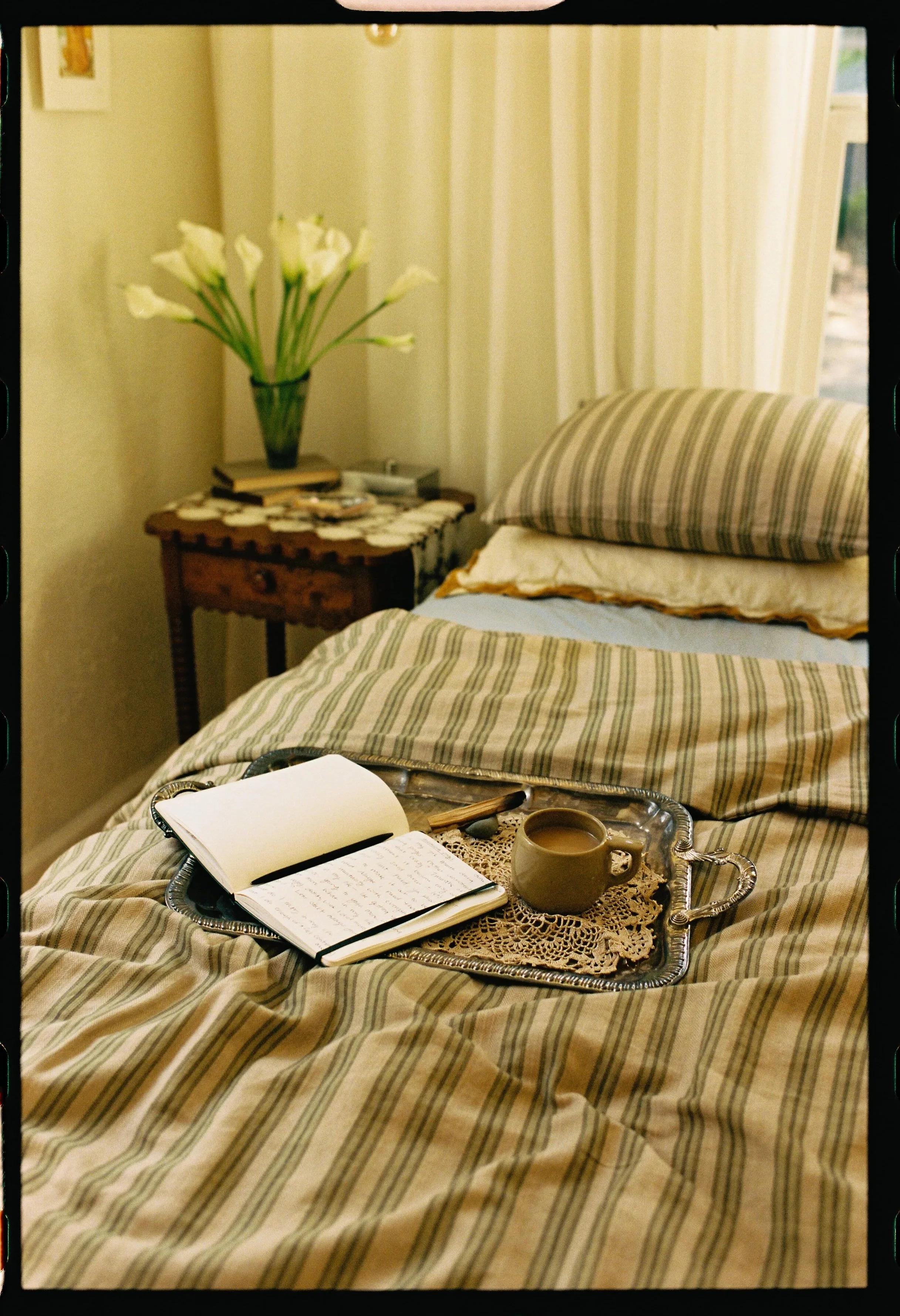 A cozy bedroom scene featuring a bed with striped bedding, a tray with a notebook, pen, and a coffee mug, and a bedside table with a vase of white calla lilies, books, and small items, near a window with light-colored curtains.