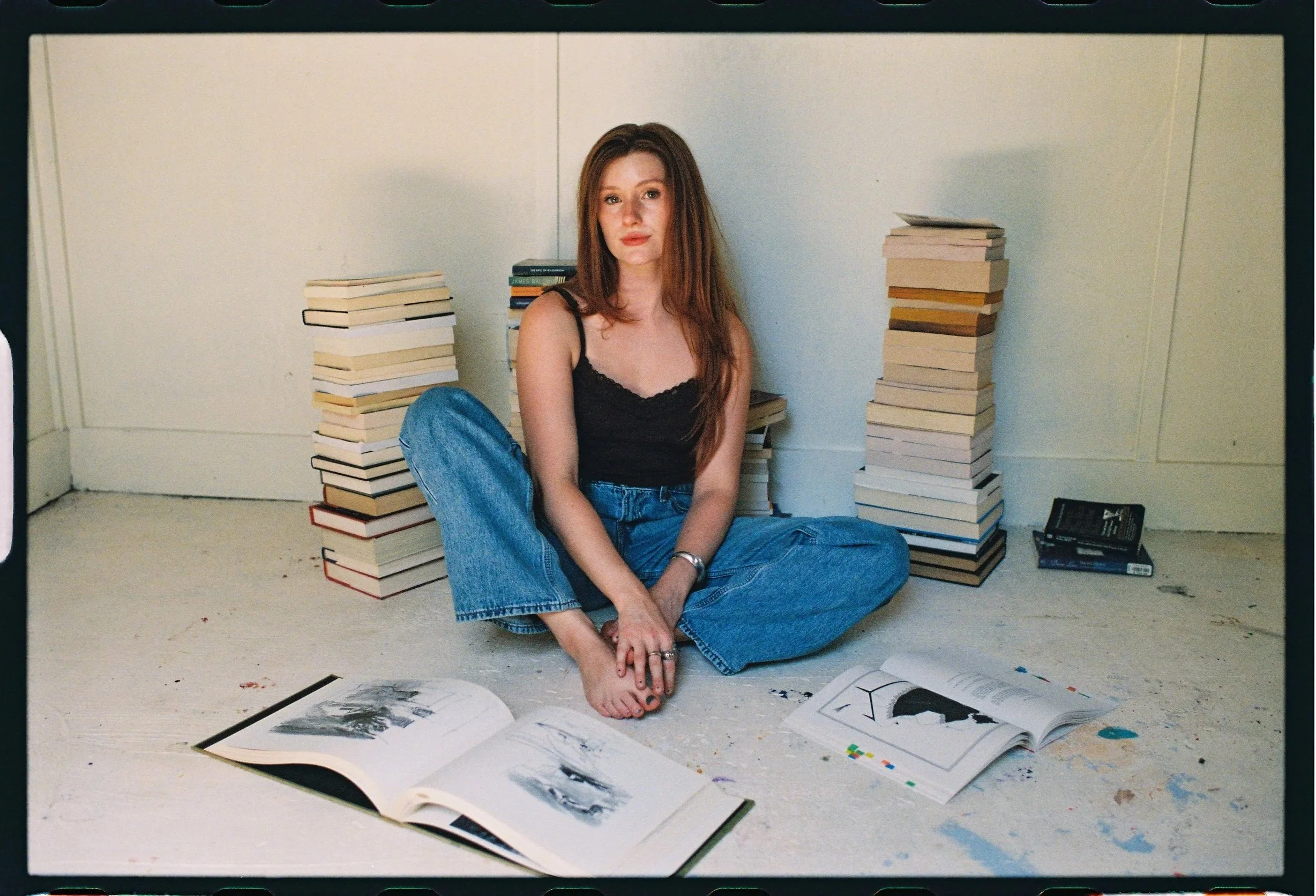 A young woman with red hair sitting on the floor, surrounded by stacks of books and open art books displaying black and white images.