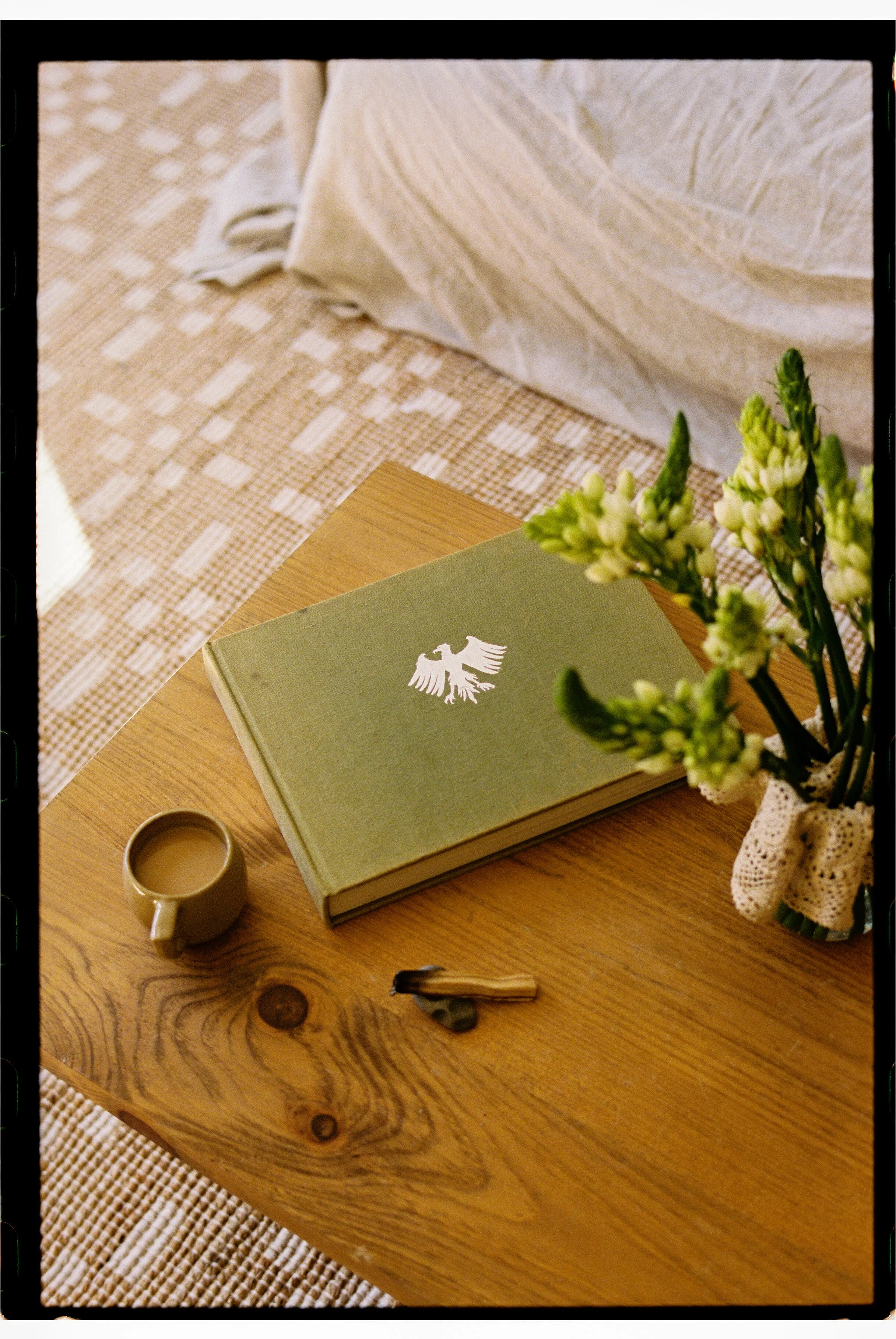 A wooden table with a green book, a beige coffee mug filled with coffee, a flower bouquet, and a pair of glasses, with a bed in the background.