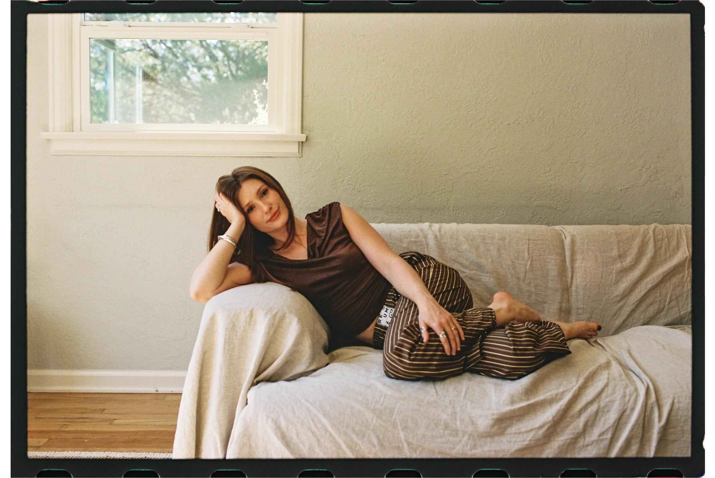 A woman lying on a beige sofa in a room with a window showing trees outside. She is resting her head on her left hand and looking at the camera. She has brown hair and is wearing a dark satin top and striped pants.