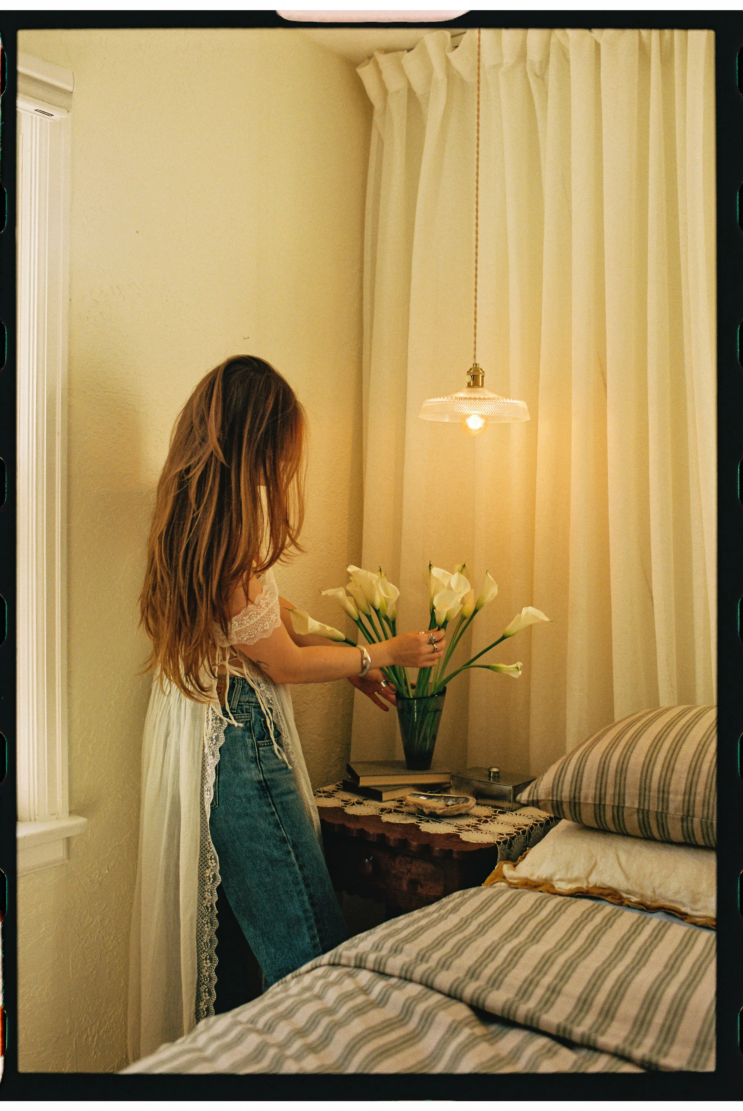 A woman with long, wavy hair wearing a lace top and jeans is arranging white calla lilies in a tall dark green vase on a side table next to a bed in a bedroom with beige walls, cream curtains, and a ceiling light fixture.
