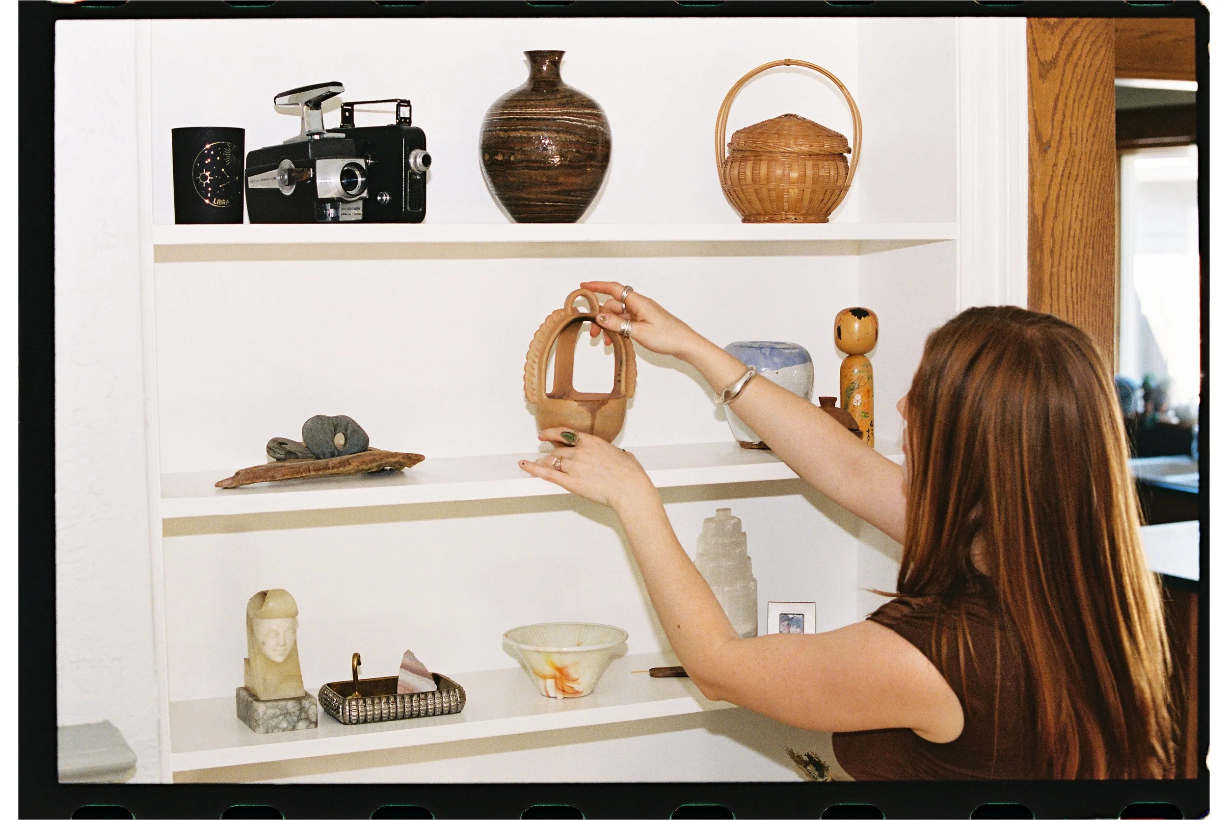 A person with brown hair arranging decorative items on a white shelf, including pottery, a sculpture, and small objects.