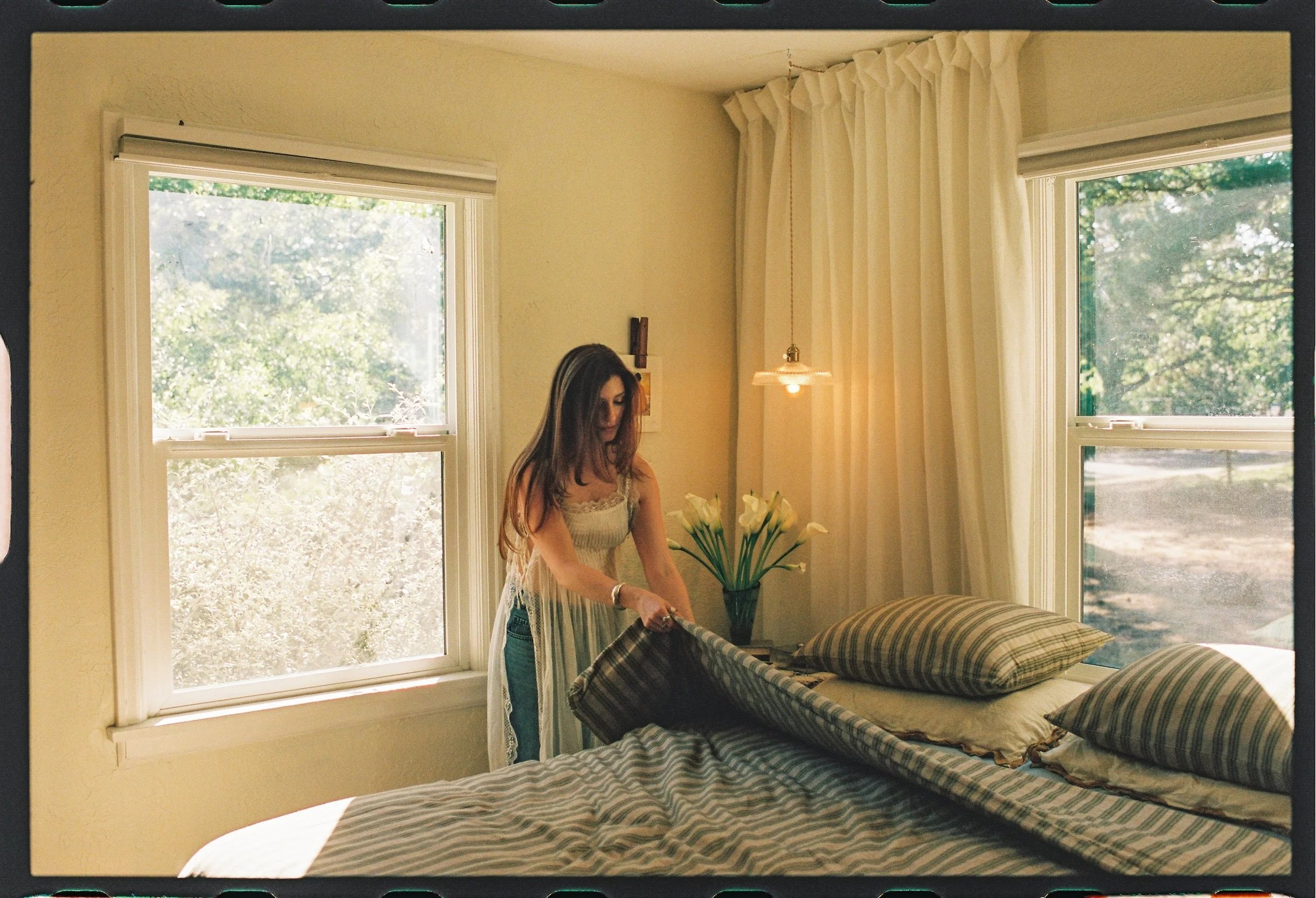 A woman with long dark hair wearing a beige sleeveless top and blue jeans stands in a bedroom near two large windows with white curtains, holding a blanket or piece of fabric, with a bed covered in a striped bedspread and some pillows in the foreground, and a vase of white calla lilies on a nightstand.