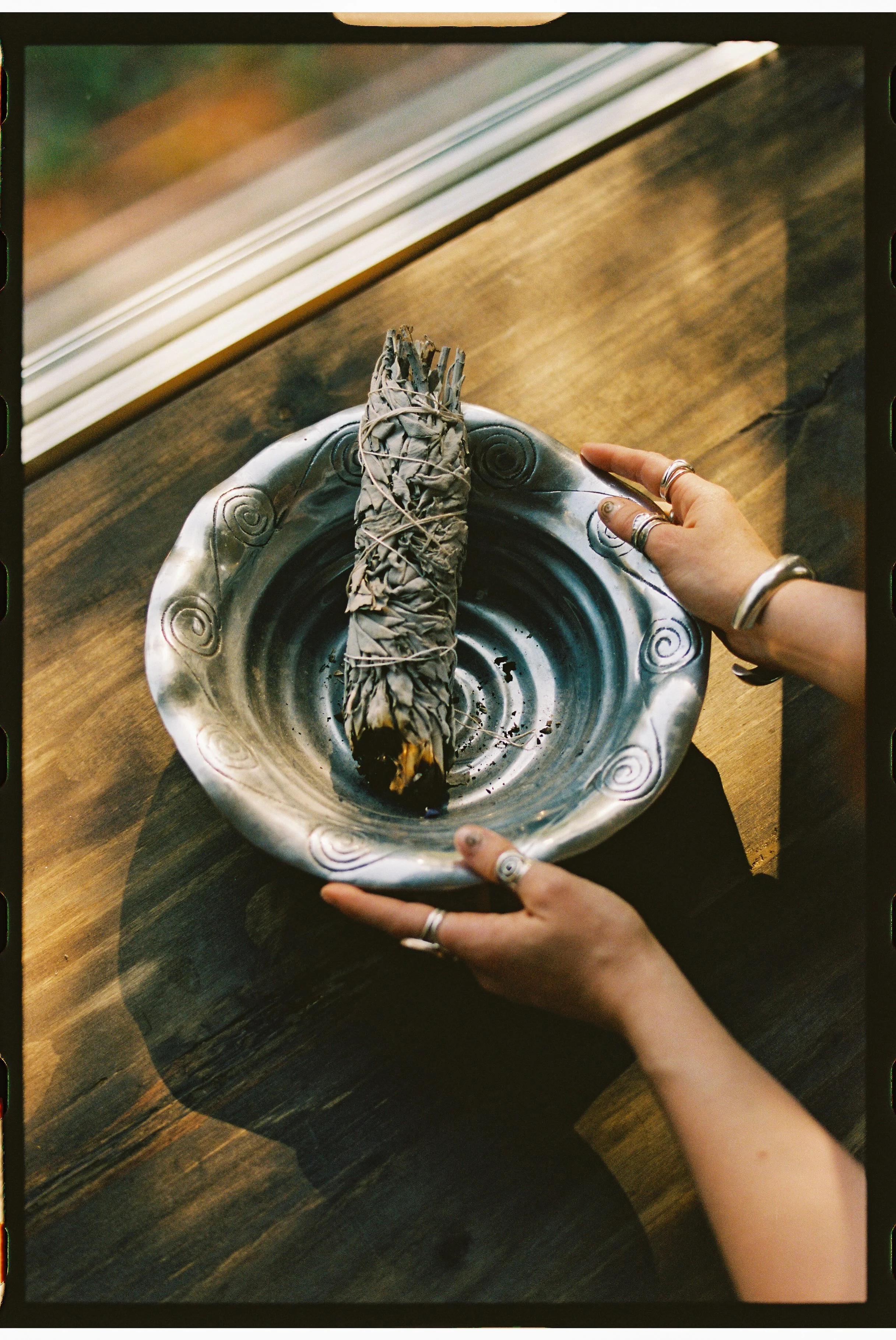 A person with rings on their fingers holds a decorated mortar pestle with dried sage and string, over a metal tray with spiral designs, on a wooden table.