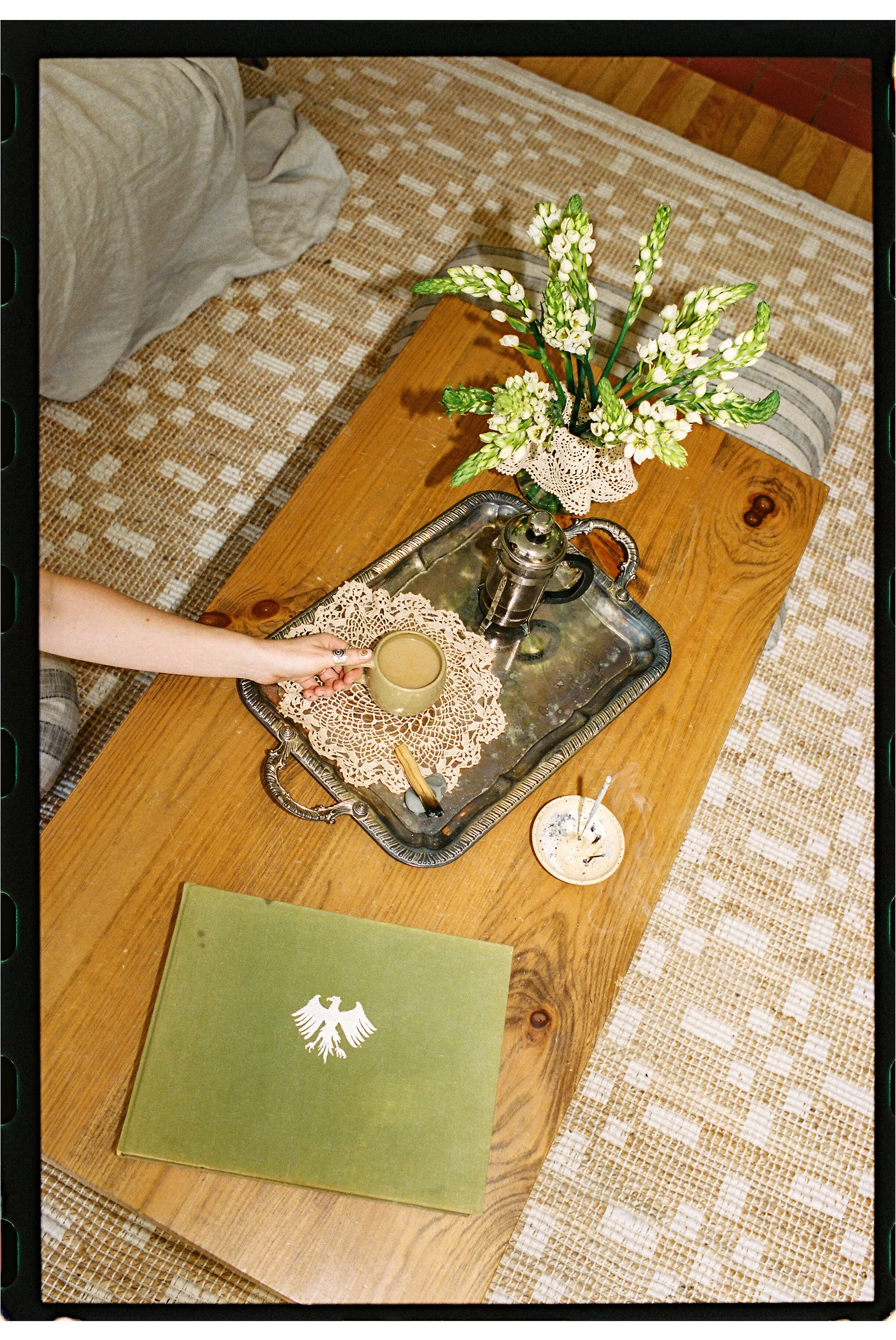 A wooden dining table with a metal tray, a white lace doily, a green book with a white eagle emblem, a small candle, a silver coffee pot, and a green vase with white flowers. There is a beige and white checkered rug underneath the table and a cushioned bench with beige and striped upholstery next to it.