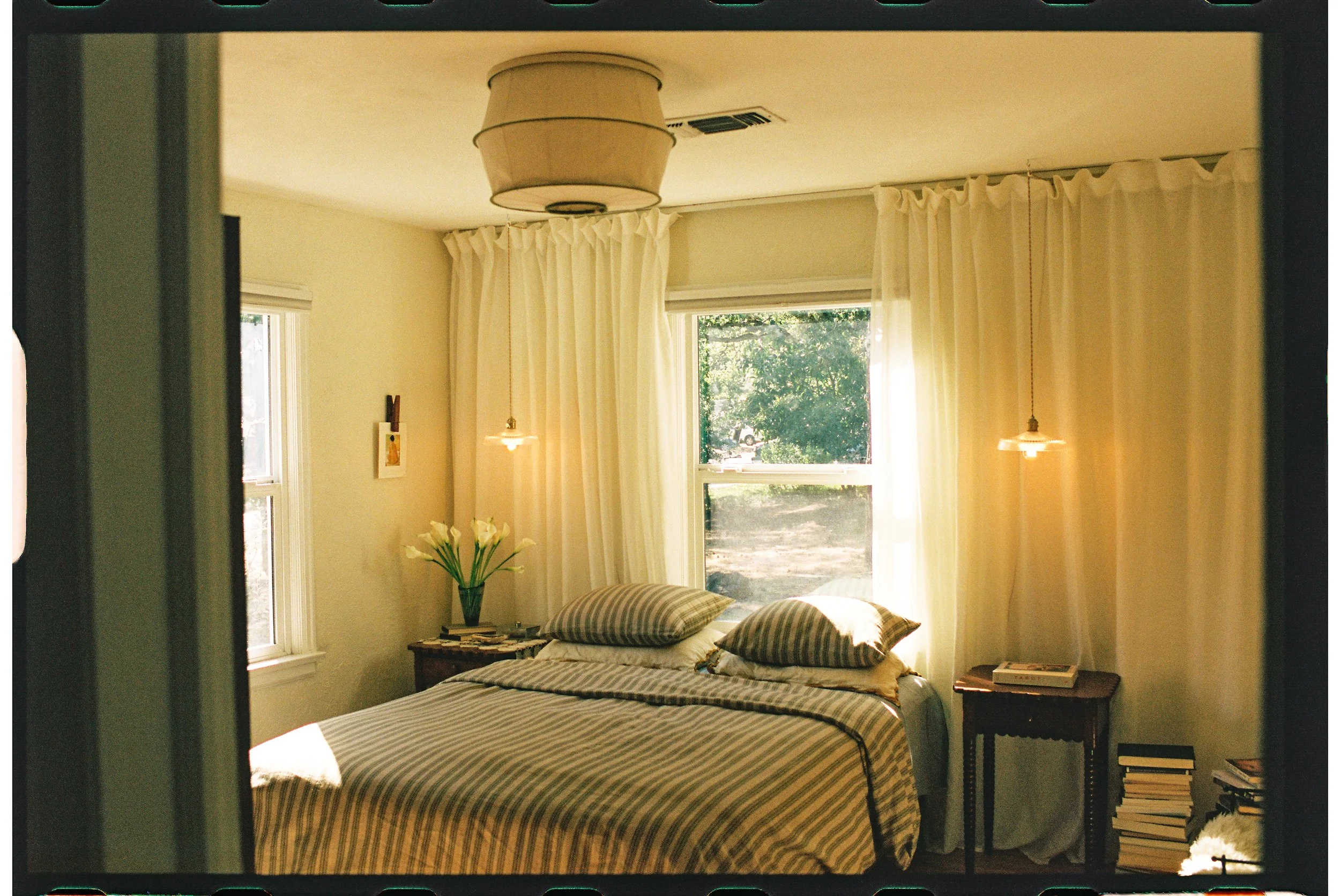 A cozy bedroom with a bed covered in a striped bedspread, two pillows, a small wooden side table with a vase of white flowers, and cream curtains, with sunlight streaming through the windows.