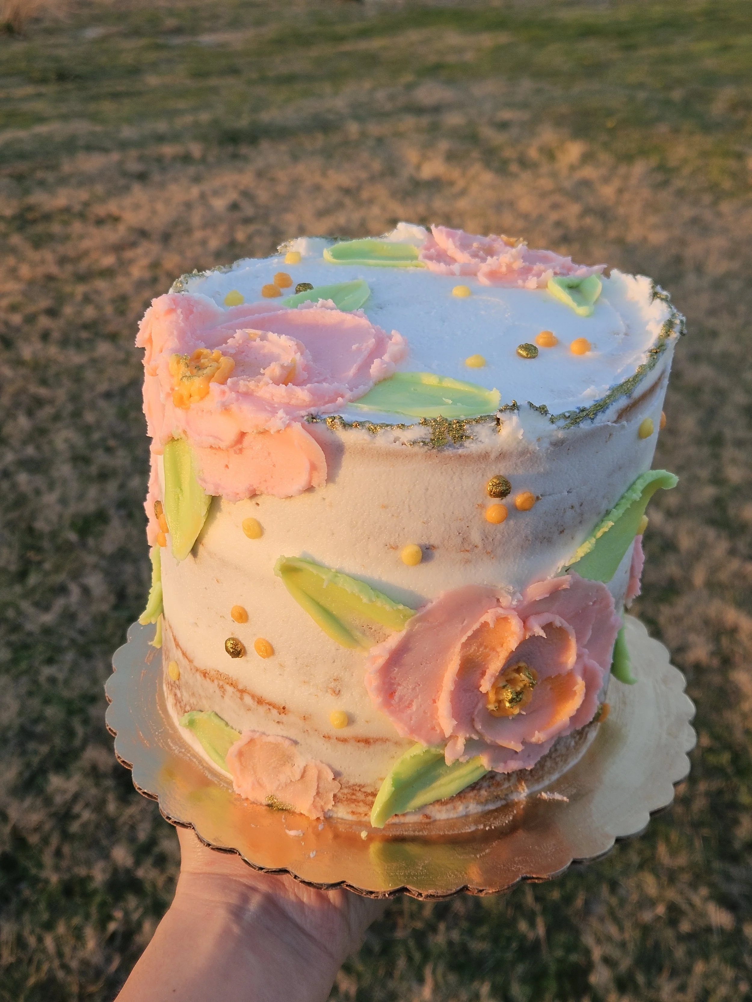 A hand holding a decorated cake outdoors with grass in the background. The cake has pastel-colored frosting, flower-like decorations, and edible glitter.
