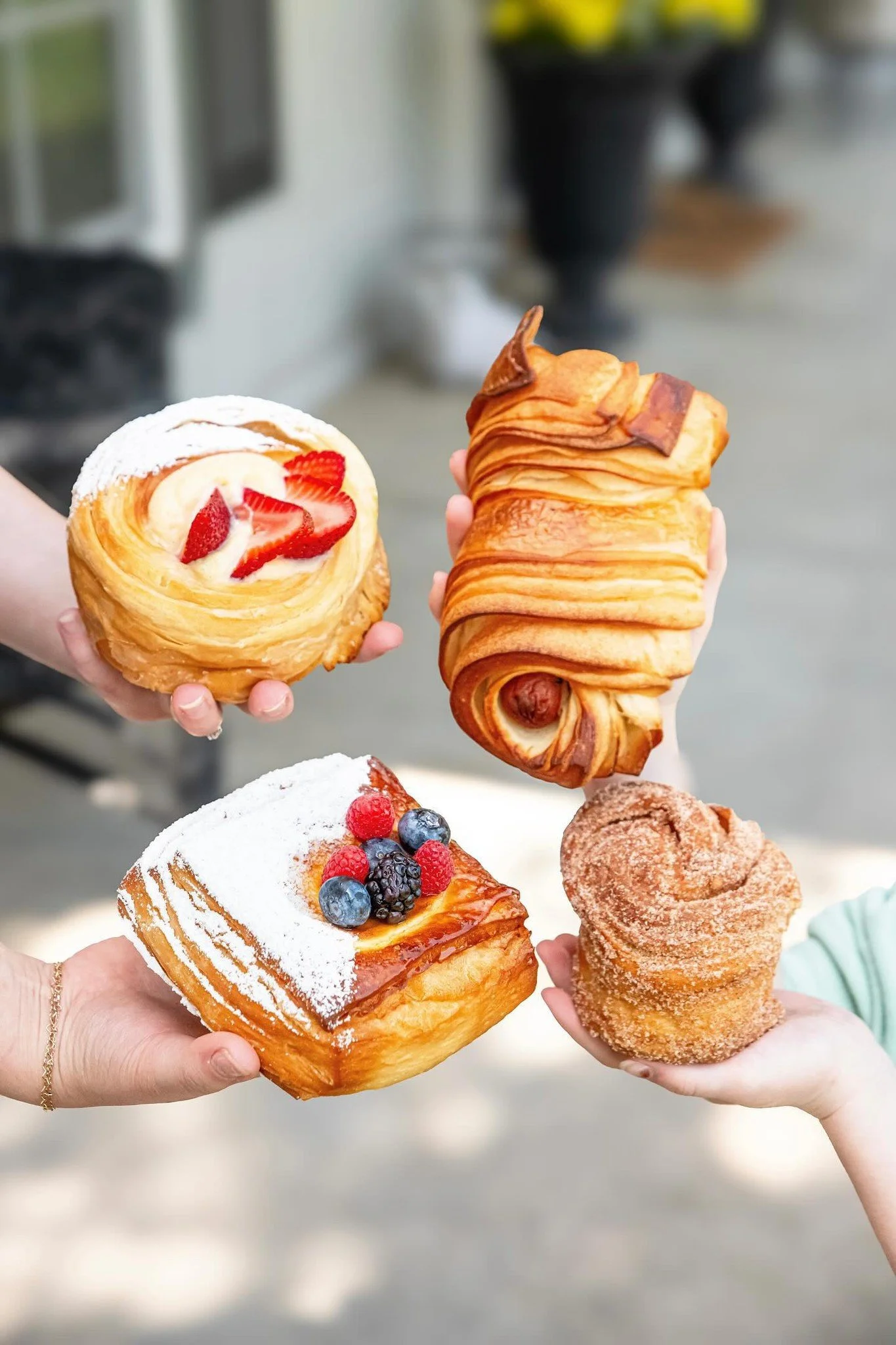 Hands holding various pastries, including a croissant with strawberries, a sweet roll, a Danish with berries, and other baked goods, against a blurred background.