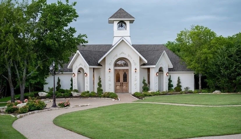 White chapel with wooden doors, surrounded by green grass and trees, with a paved walkway leading to the entrance.