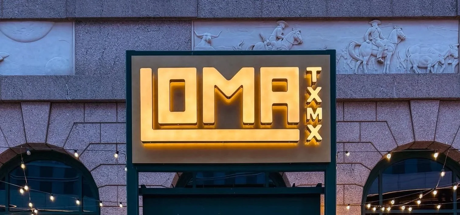 Sign for Omaha, Nebraska with illuminated yellow letters, display with sculptures on the building wall behind, and string lights in front.