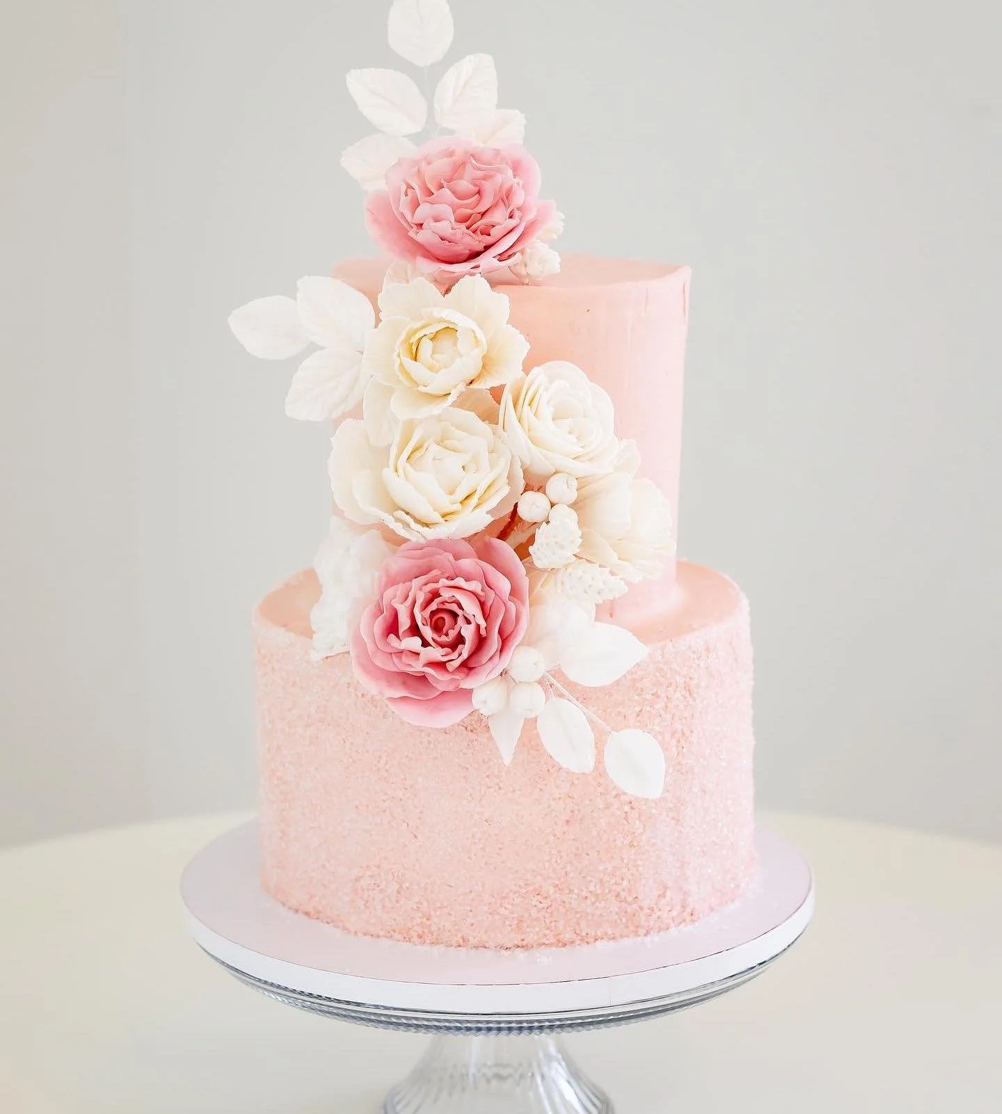 A two-tier pink cake decorated with pink and white flowers, including roses, with a white leaf design, displayed on a glass cake stand.