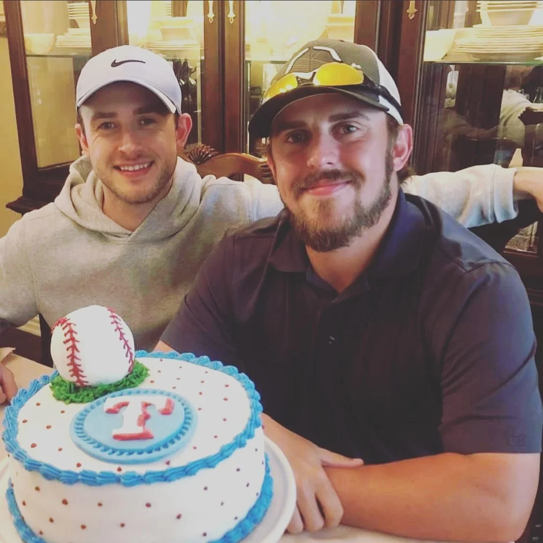 Two men sitting at a dining table, smiling, with a baseball-themed birthday cake in front of them. The cake is decorated with a baseball and a red football-shaped 'T' emblem.