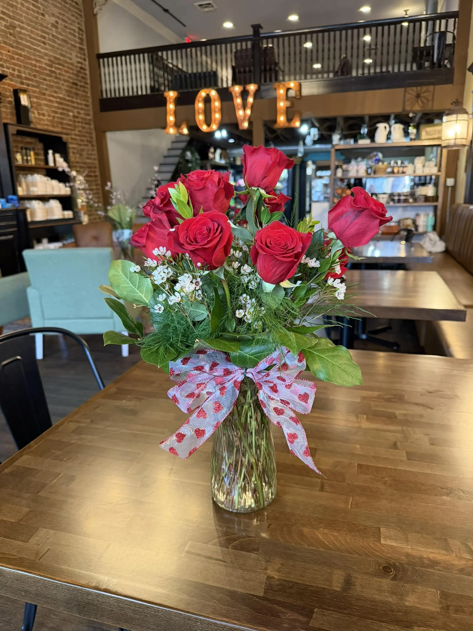 A bouquet of red roses and white filler flowers in a clear glass vase with a red and white patterned bow, placed on a wooden table inside a cafe or restaurant with a brick wall and wooden shelving in the background.