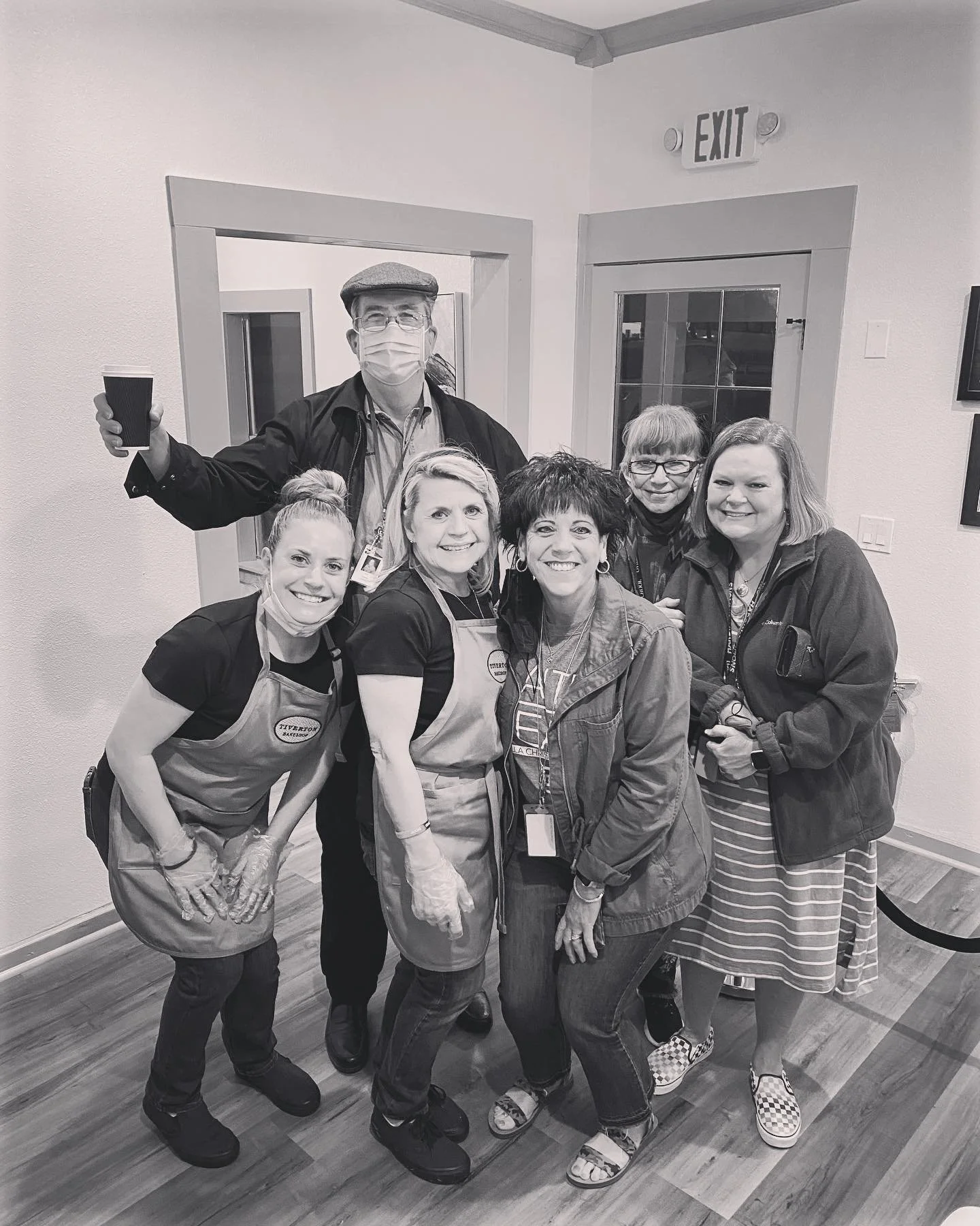 Group of six women and one man in a room, smiling for the camera. The man is wearing a face mask, glasses, a flat cap, a dark jacket, and is holding a paper cup. The women are wearing aprons, casual clothes, and some have masks pulled down. The room has an EXIT sign above a door, and framed pictures on the wall.