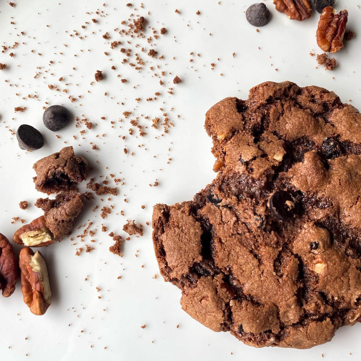 Chocolate chip cookie with scattered crumbs, chocolate chips, and pecan pieces on a white surface.