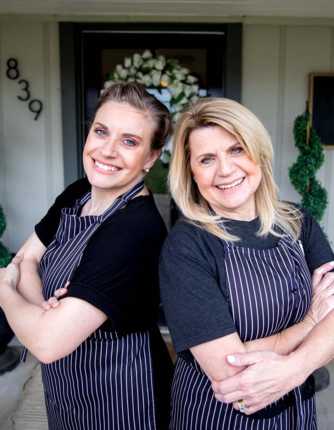 Two smiling women in black shirts and striped aprons standing outside in front of a house with a floral wreath on the door.