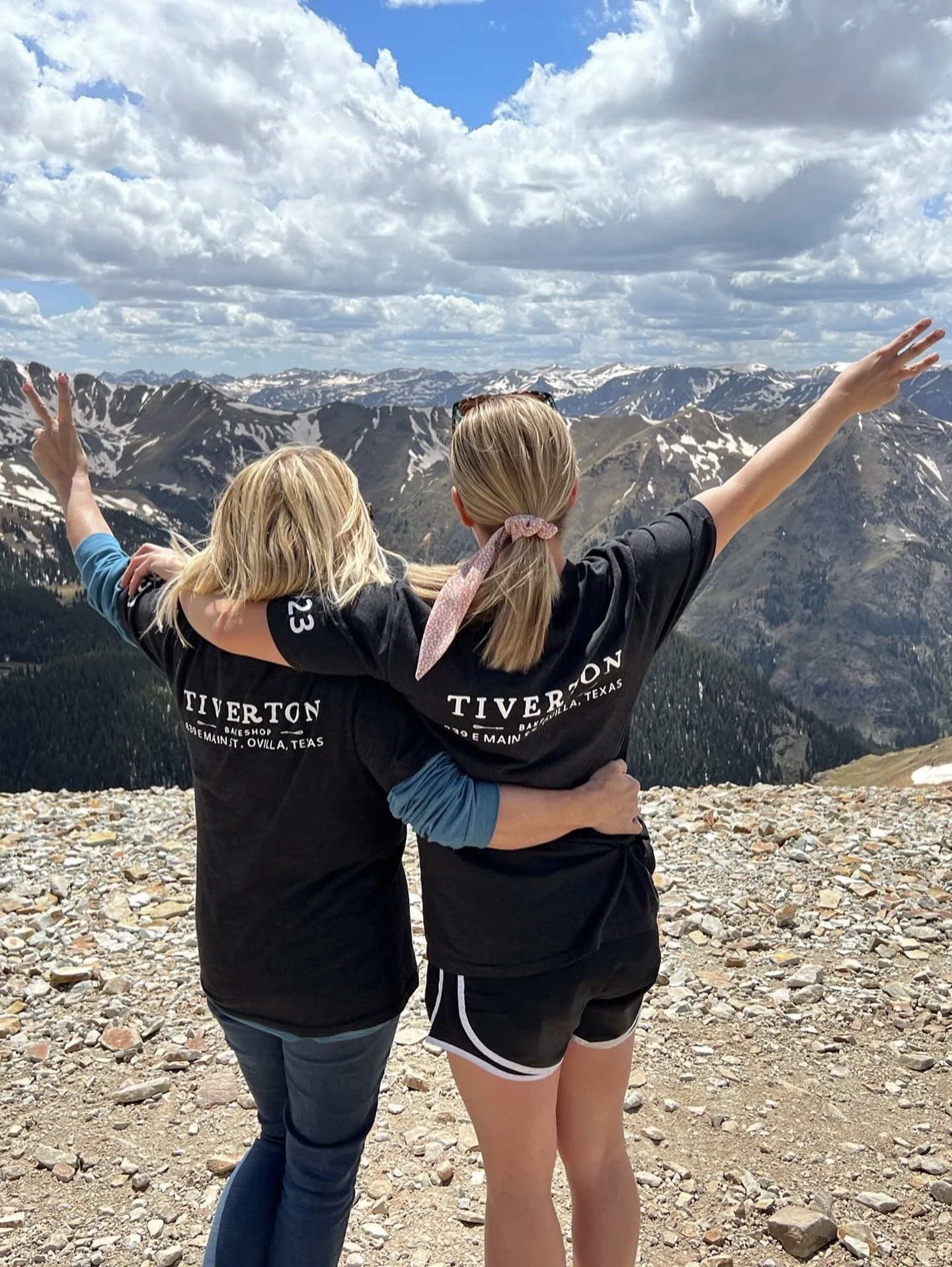 Two women embrace overlooking snow-capped mountains and a partly cloudy sky, with one woman making a peace sign with her hand.