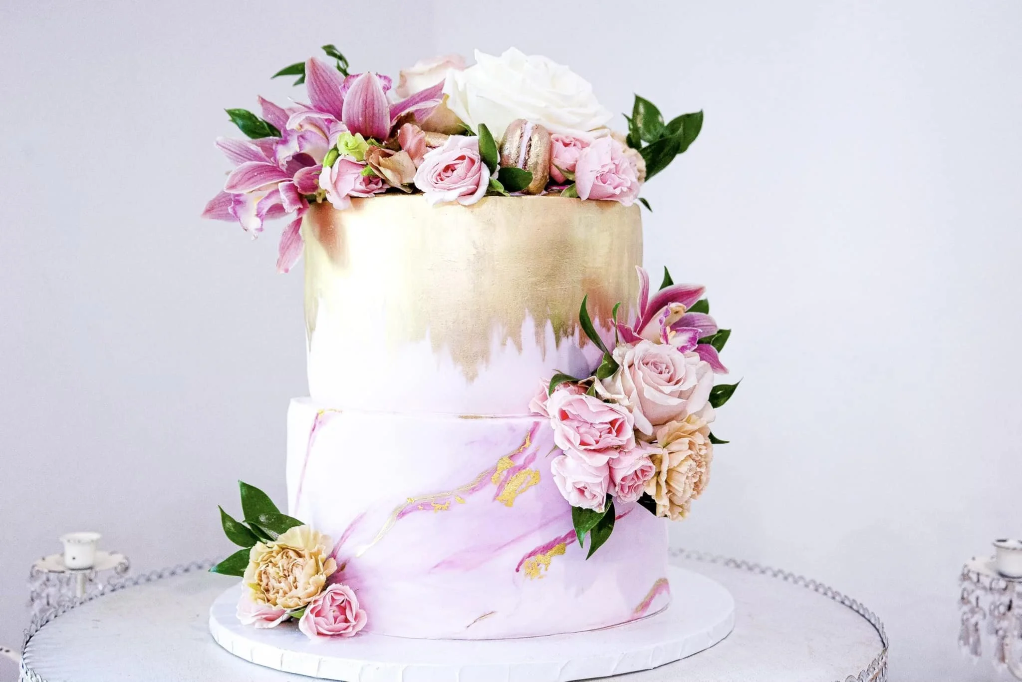 A two-tiered wedding cake decorated with pink and white flowers, including roses and lilies, with a white and pink marbled frosting design, placed on a white cake stand.