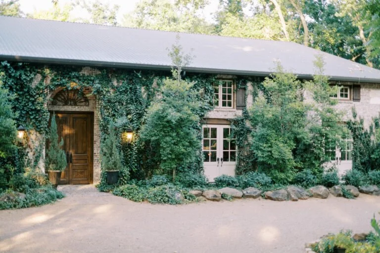 A house covered with green ivy and trees, with a wooden front door and multiple windows, surrounded by a stone-lined garden bed and a gravel driveway.