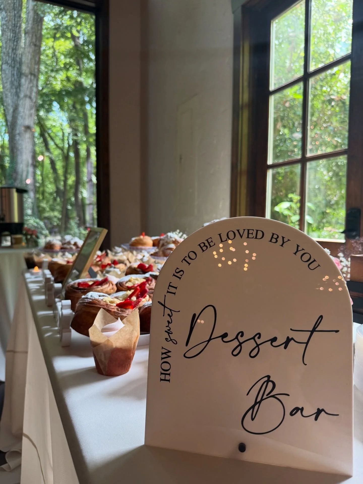 A table of desserts indoors near large windows with trees outside. The desserts include cupcakes and other pastries. A white sign with black cursive text reads, 'How sweet it is to be loved by you' and 'Dessert Bar.'