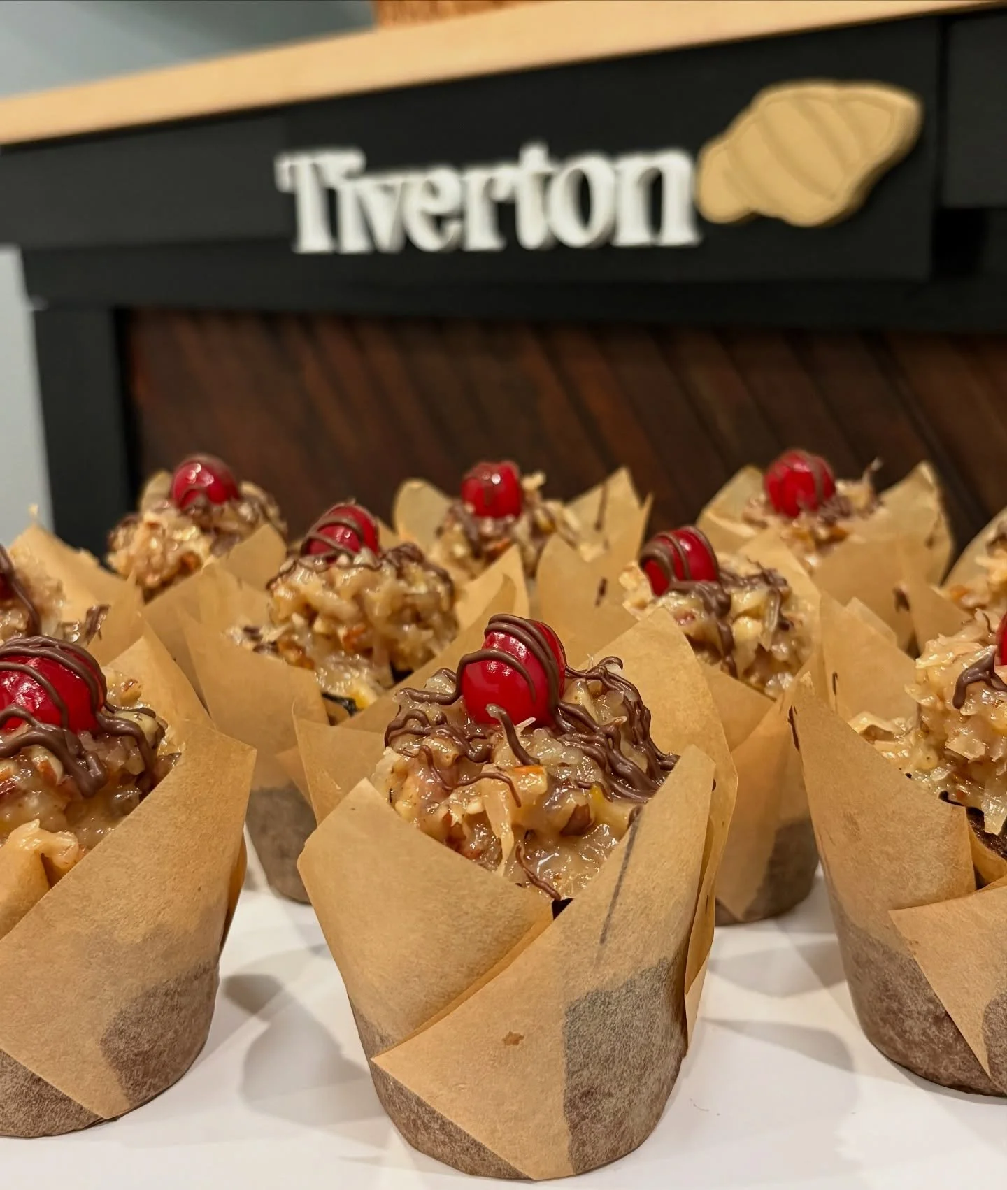 Cupcakes topped with chocolate drizzle, chopped nuts, and a cherry in paper wrappers in front of a Tverton bakery sign with a croissant logo.