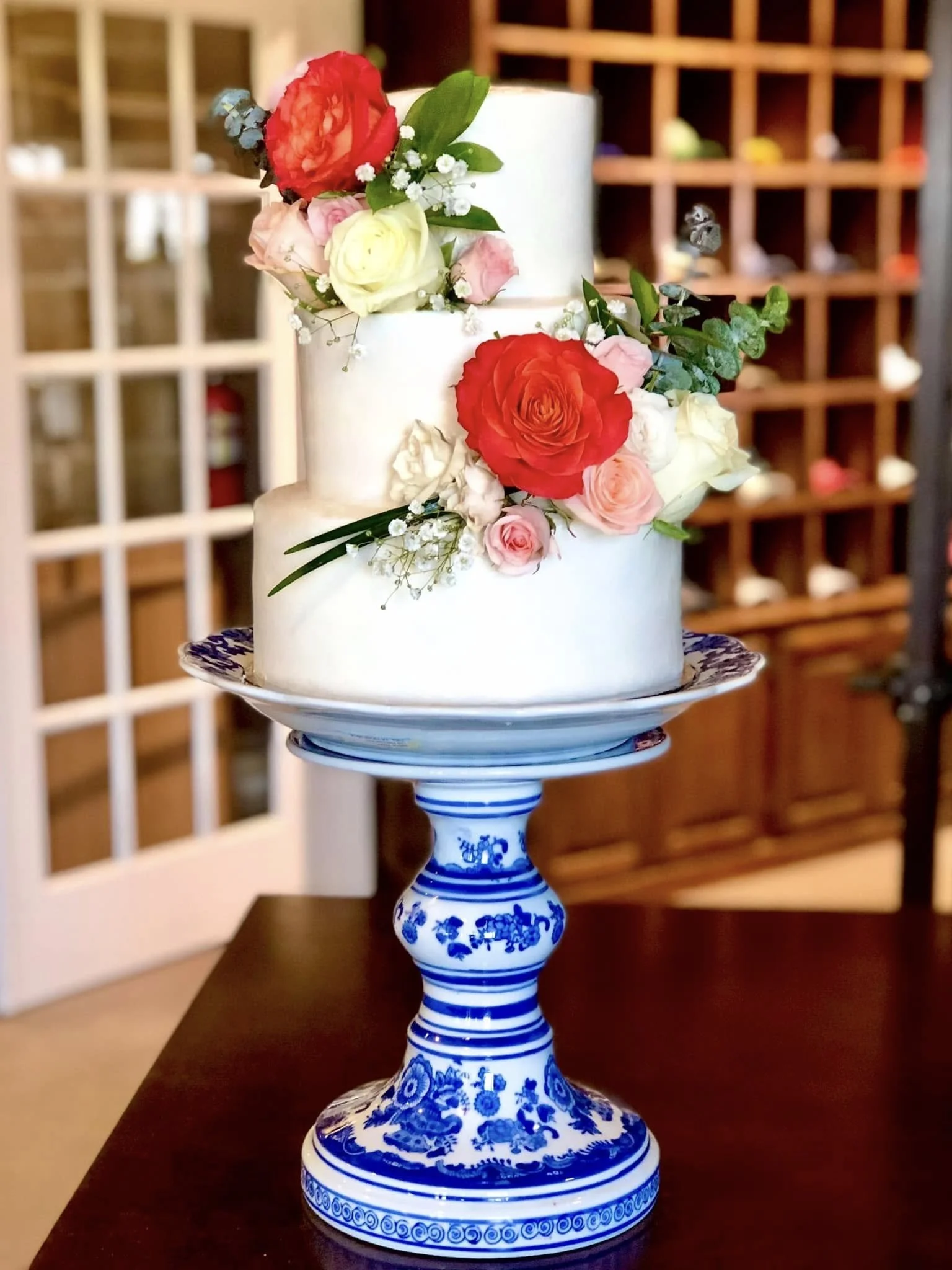A two-tiered white wedding cake decorated with an assortment of pink, red, yellow, and white flowers and greenery, placed on a blue and white ceramic cake stand.