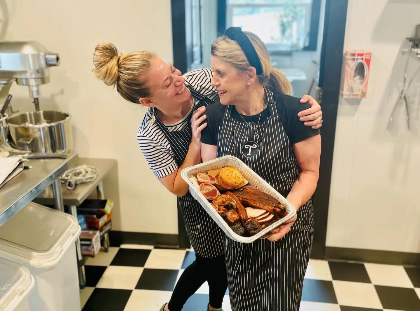 Two women in striped aprons sharing a joyful moment in a kitchen, one holding a tray of cooked barbecue meat, side dishes, and a baked item.