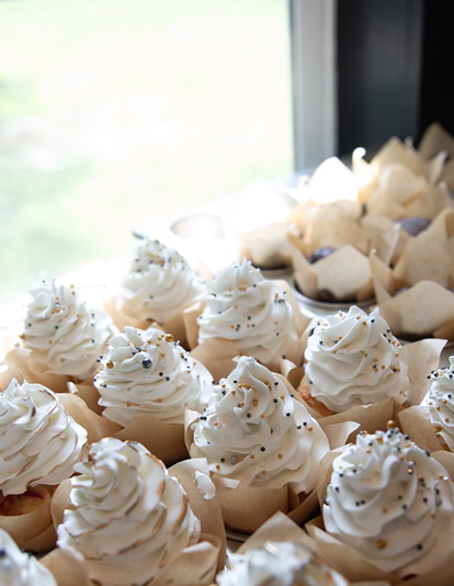 Rows of cupcakes with white frosting and silver sprinkles inside paper cups near a window.