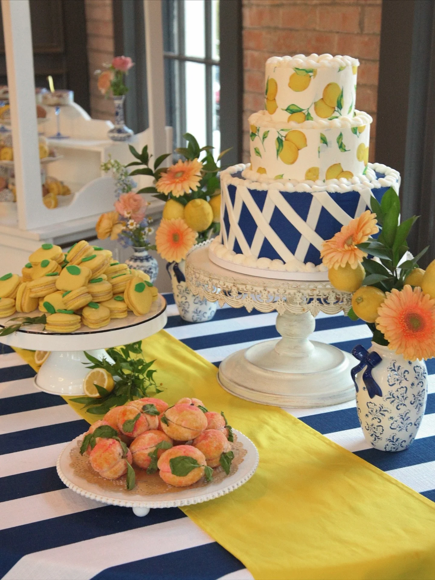 A dessert table with a three-tier lemon-themed cake, lemon decor, and flowers, with plates of cookies and other treats on a blue and white striped tablecloth.
